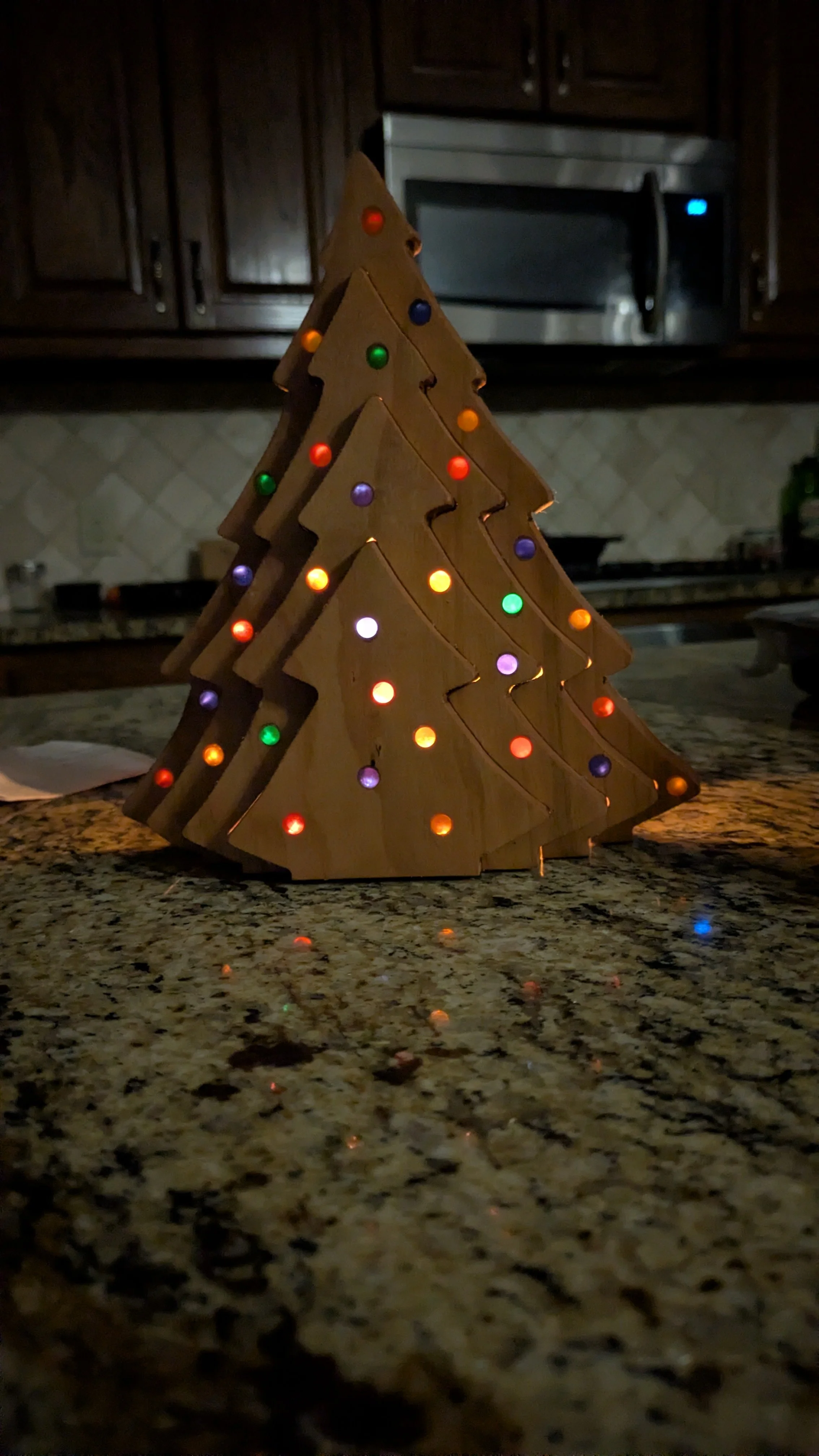 A wooden Christmas tree decoration with colorful lights, placed on a granite countertop in a kitchen.
