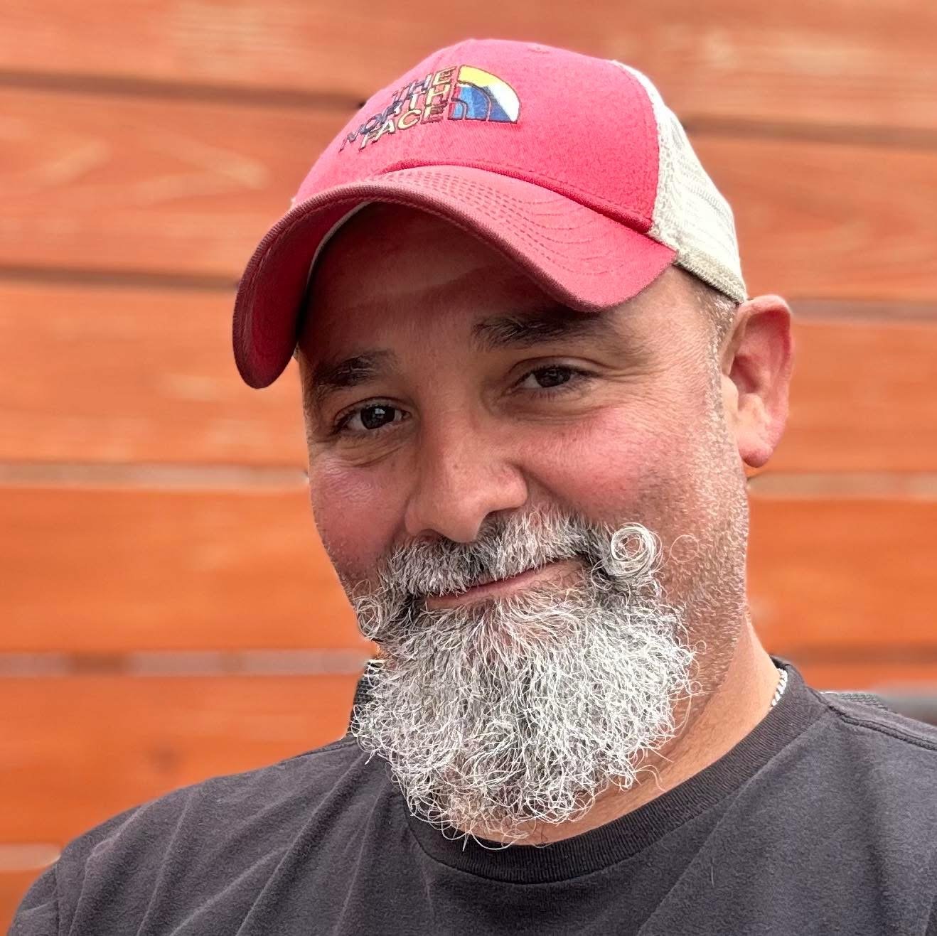 A close-up of a man with a gray beard and mustache, wearing a red and white baseball cap with The North Face logo, and a black t-shirt, standing against a wooden wall background.