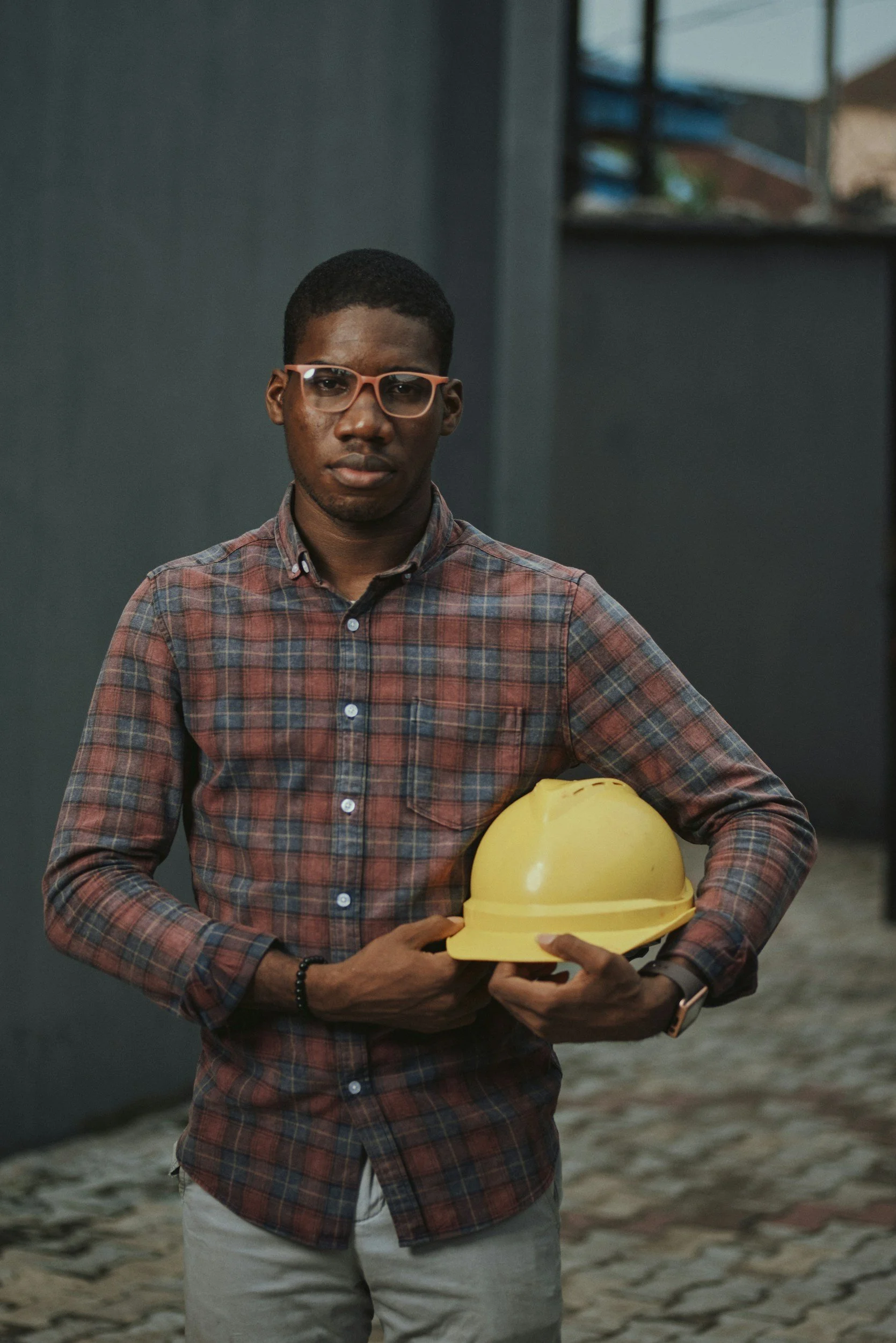 A young man wearing glasses and a plaid shirt is holding a yellow construction helmet in his right hand. He stands outdoors on a cobblestone path, with a serious expression, against a background of a dark gray wall and a building with an open window.
