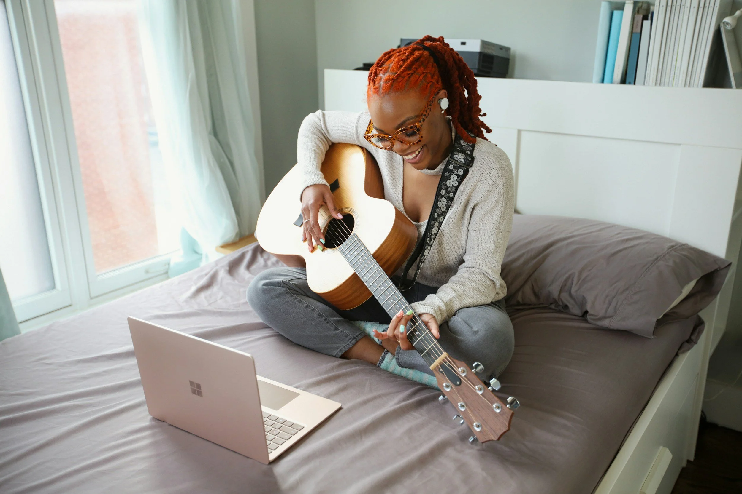 A woman with red dreadlocks wearing glasses and in-ear headphones, sitting on a bed and playing an acoustic guitar while looking at a laptop in front of her.