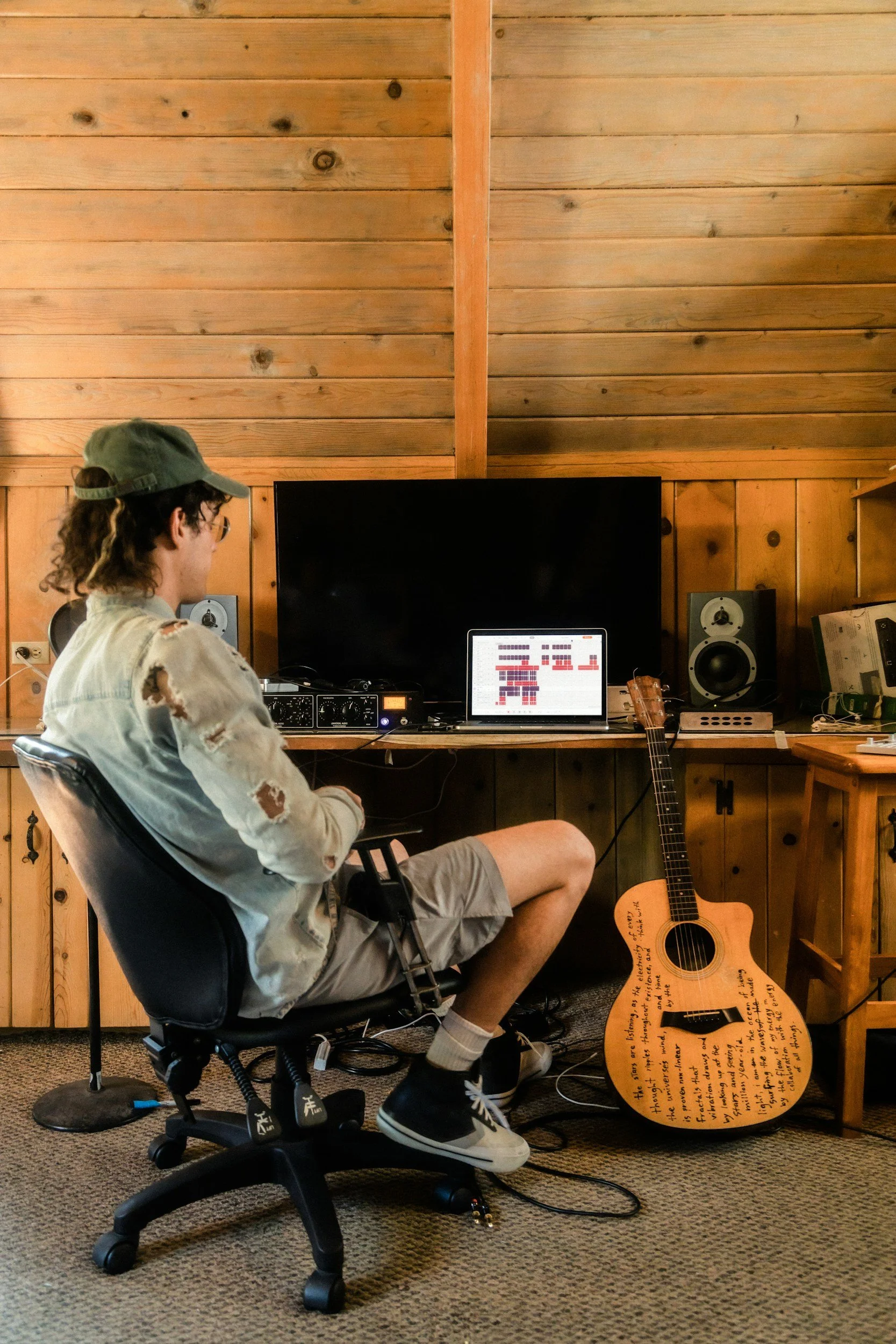 A person with curly hair, wearing glasses, a green cap, a distressed denim jacket, and shorts, sitting in a black office chair in a wooden-paneled room. They are looking at electronic music equipment and a laptop on a wooden desk, with a guitar resting on the floor beside them.