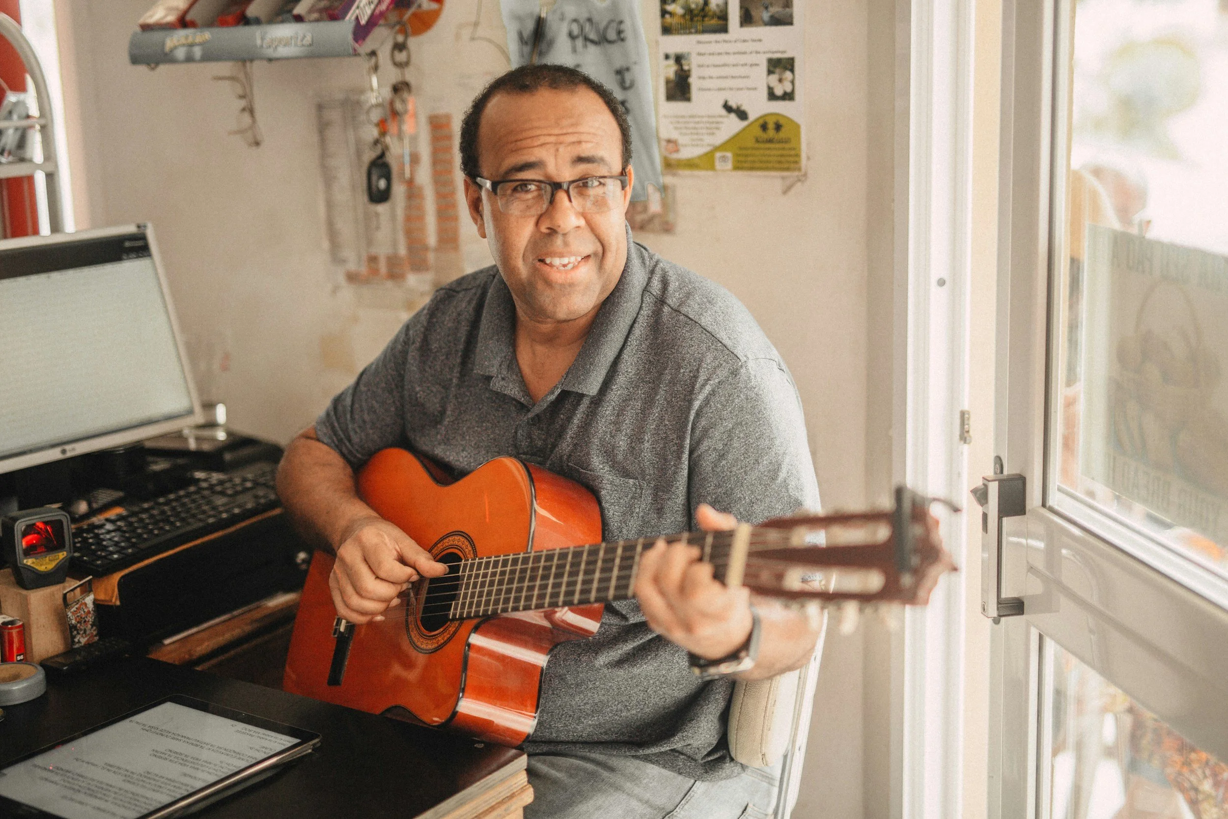 A man with glasses playing an acoustic guitar indoors, sitting at a desk with electronic equipment and papers, near a window with sunlight, in a casual environment.