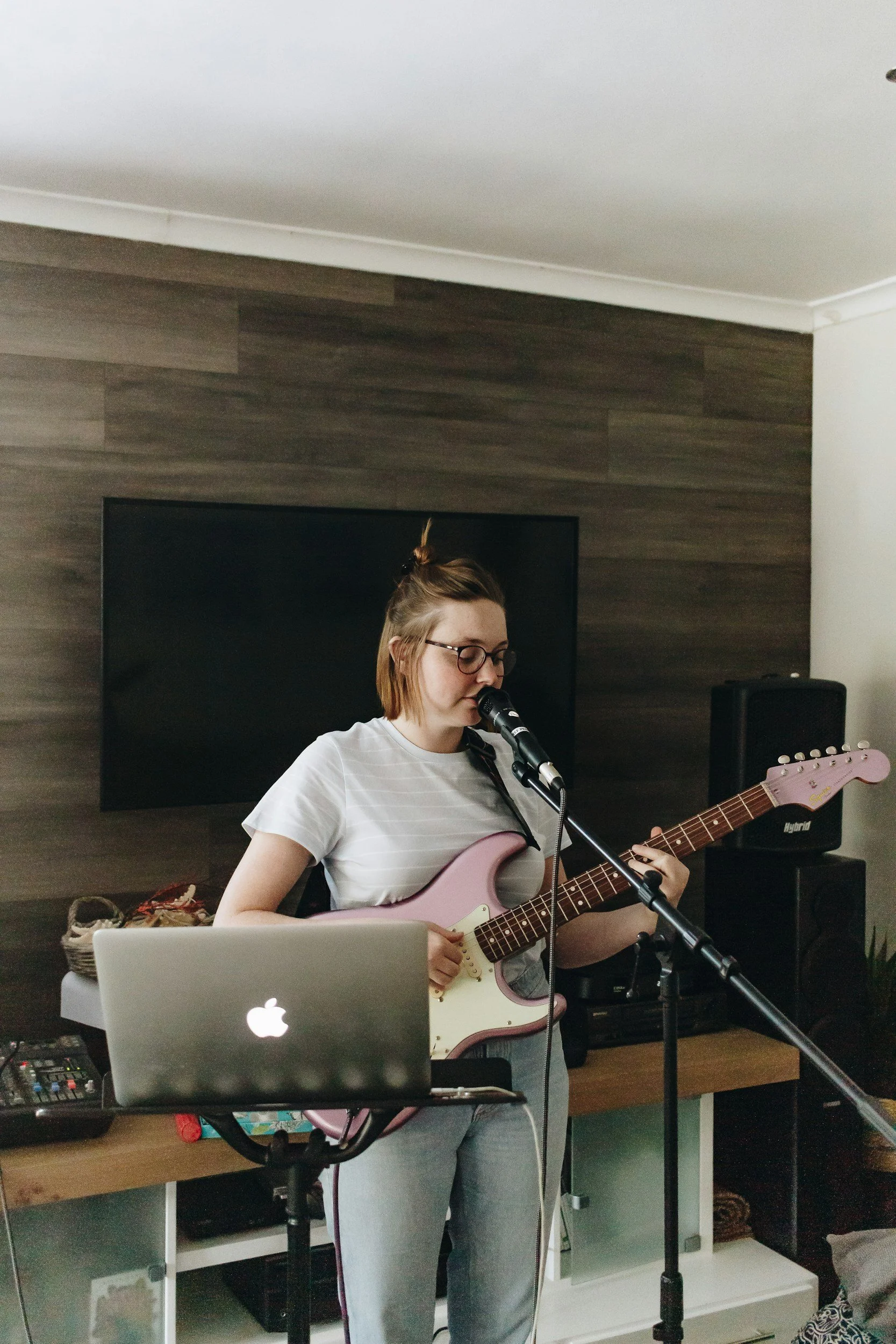 A young woman with glasses, wearing a white t-shirt and gray pants, playing an electric guitar and singing into a microphone in a home studio with dark wood paneling, a TV, a MacBook, and music equipment.