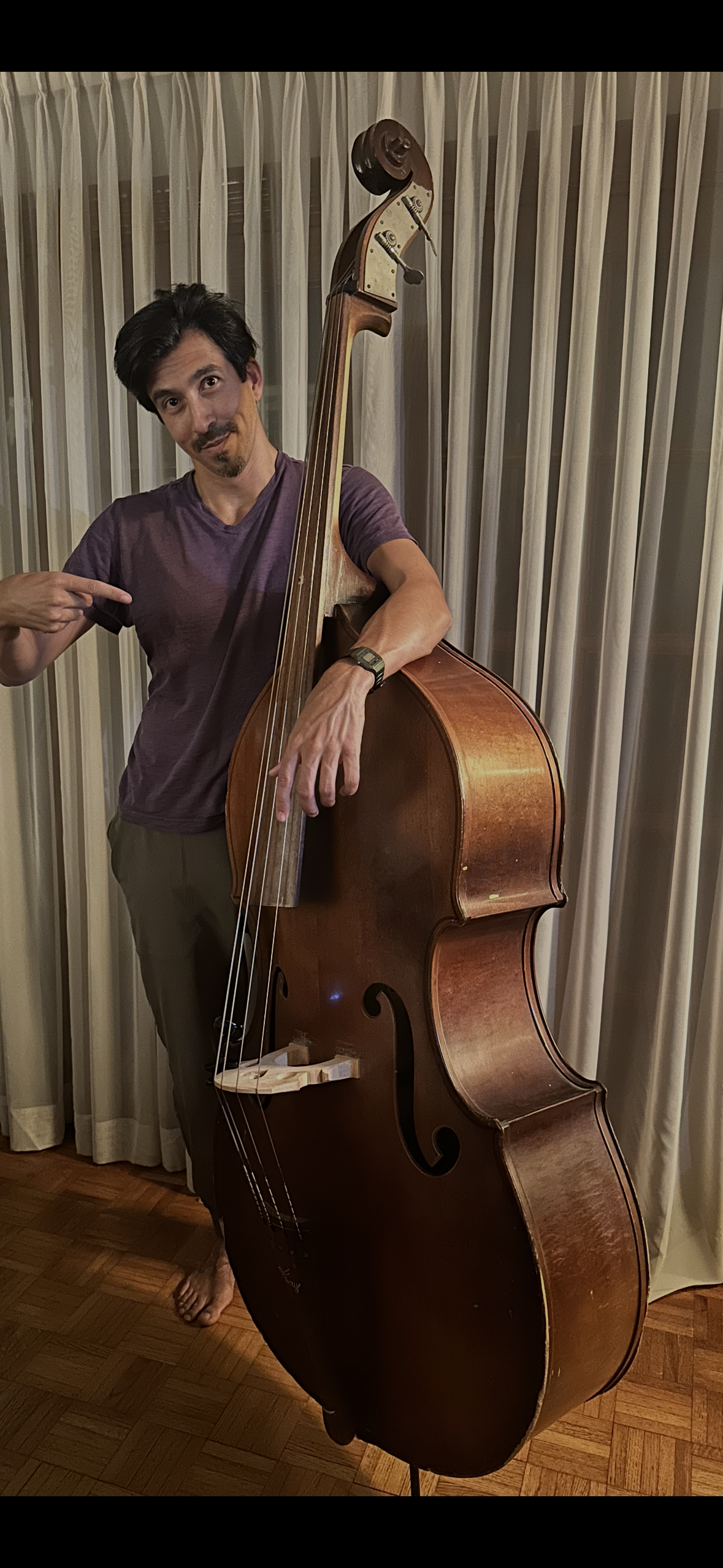 A man standing next to a large double bass, pointing at it, with a cream-colored curtain in the background.