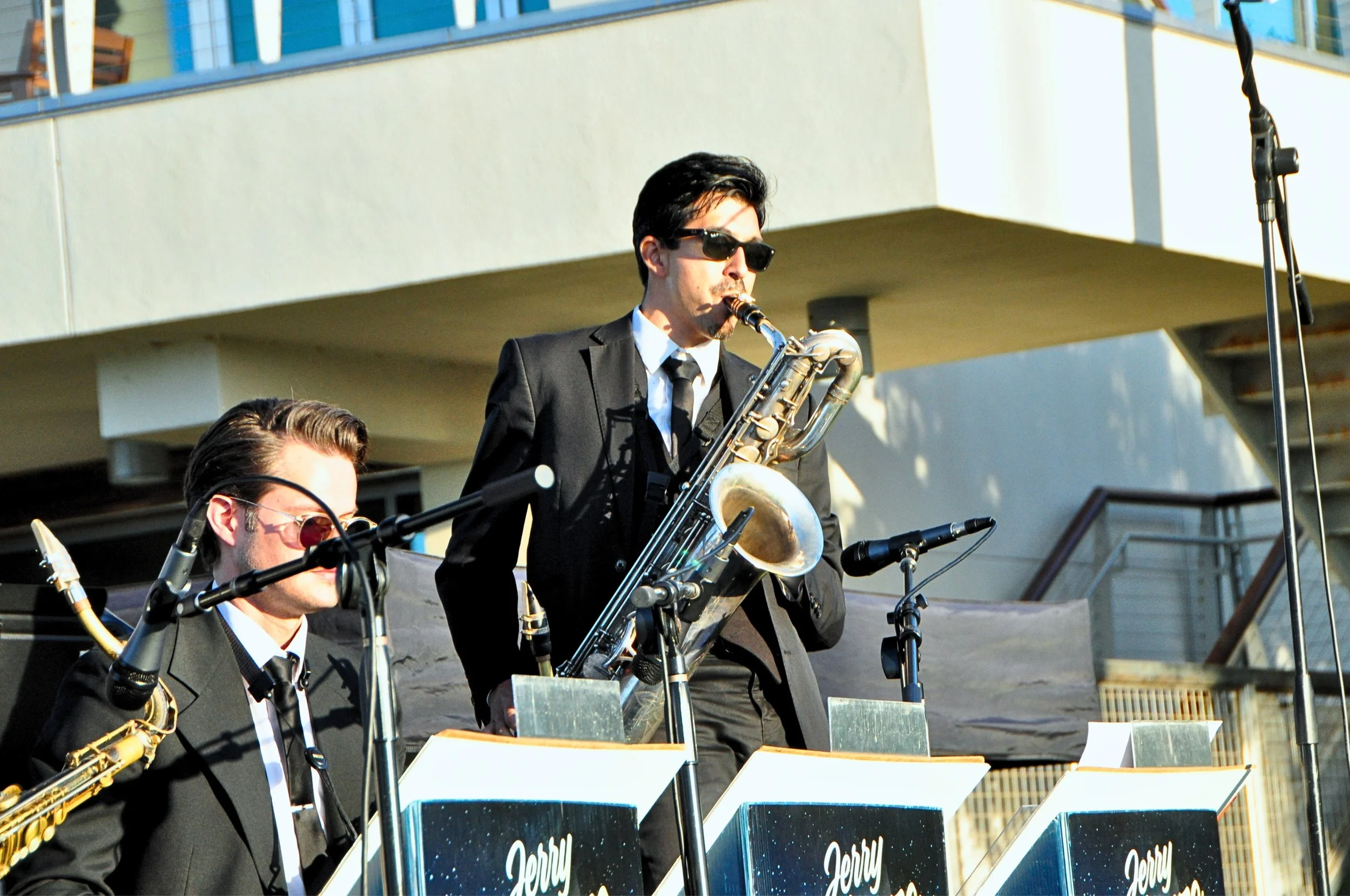 Musicians playing saxophones and other instruments during a live outdoor performance at sunset on a stage with music stands and a backdrop with starry sky design.