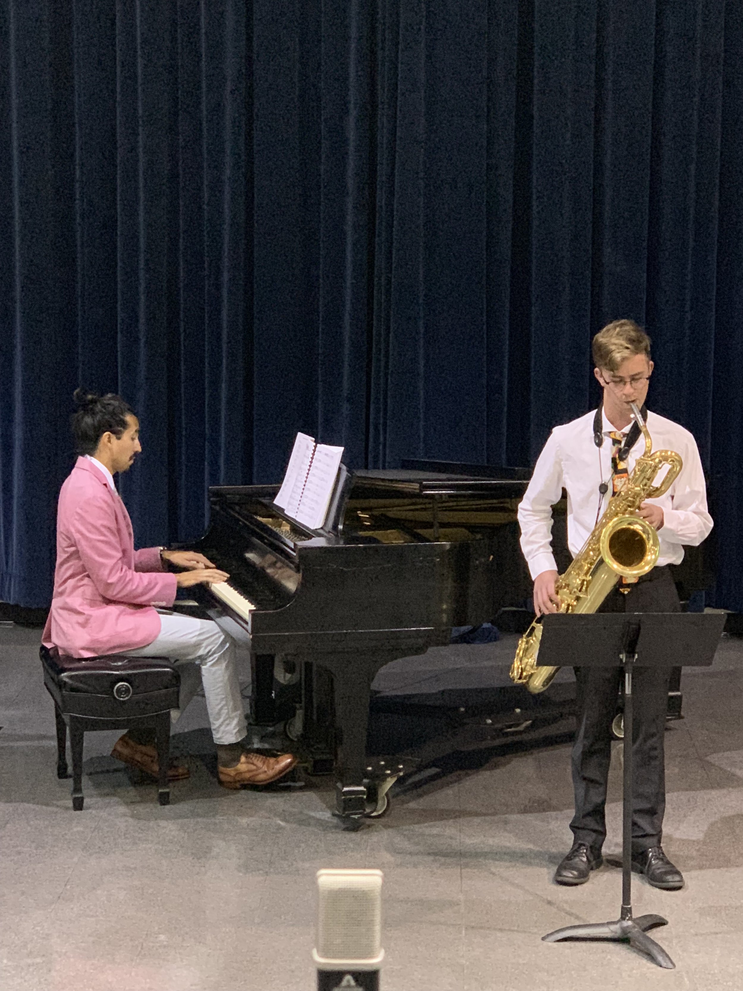 Two young musicians performing on stage: one playing piano, the other playing saxophone. They are dressed formally, with the pianist in a pink blazer and white pants, and the saxophonist in a white shirt and black pants. A music stand with sheet music is in front of the pianist, and a dark blue curtain backdrop is behind them.