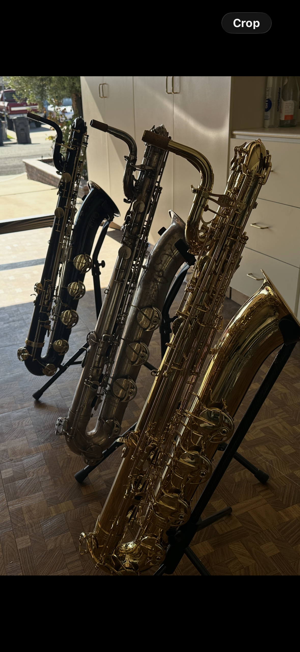 Multiple saxophones, including a gold, a silver, and a black one, standing on music stands in a room with a wooden floor and cabinets, with an outdoor scene visible through the window.