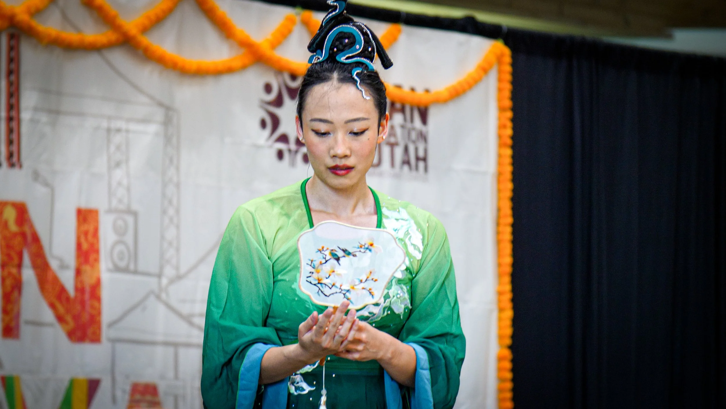 A woman dressed in a vibrant green traditional Asian costume is holding a decorative fan with an embroidered bird and flowers, standing in front of a backdrop decorated with orange marigold flowers, likely during a cultural event.