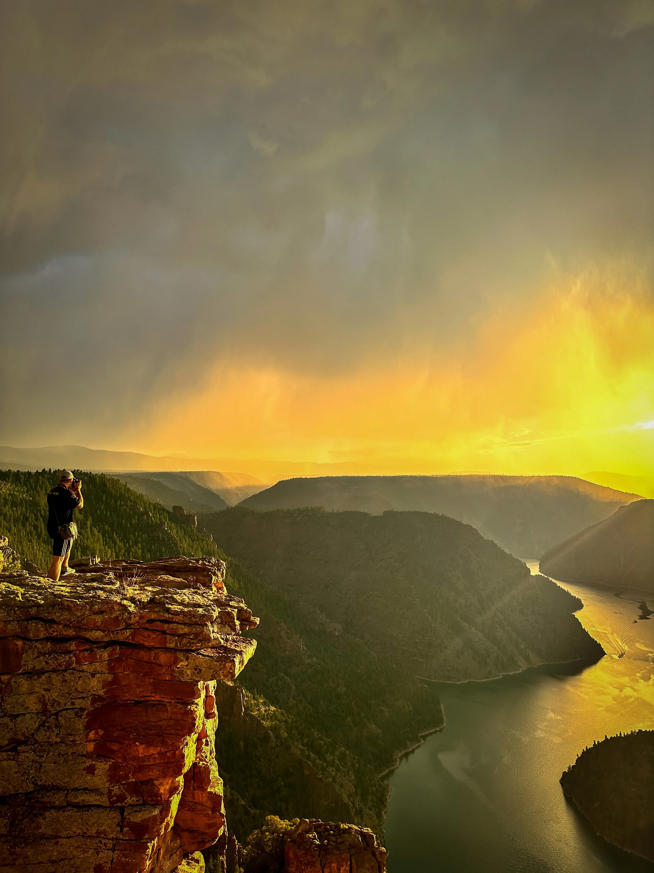 A person standing on a rocky cliff taking a photograph of a sunset over a river and canyon landscape with clouds in the sky.