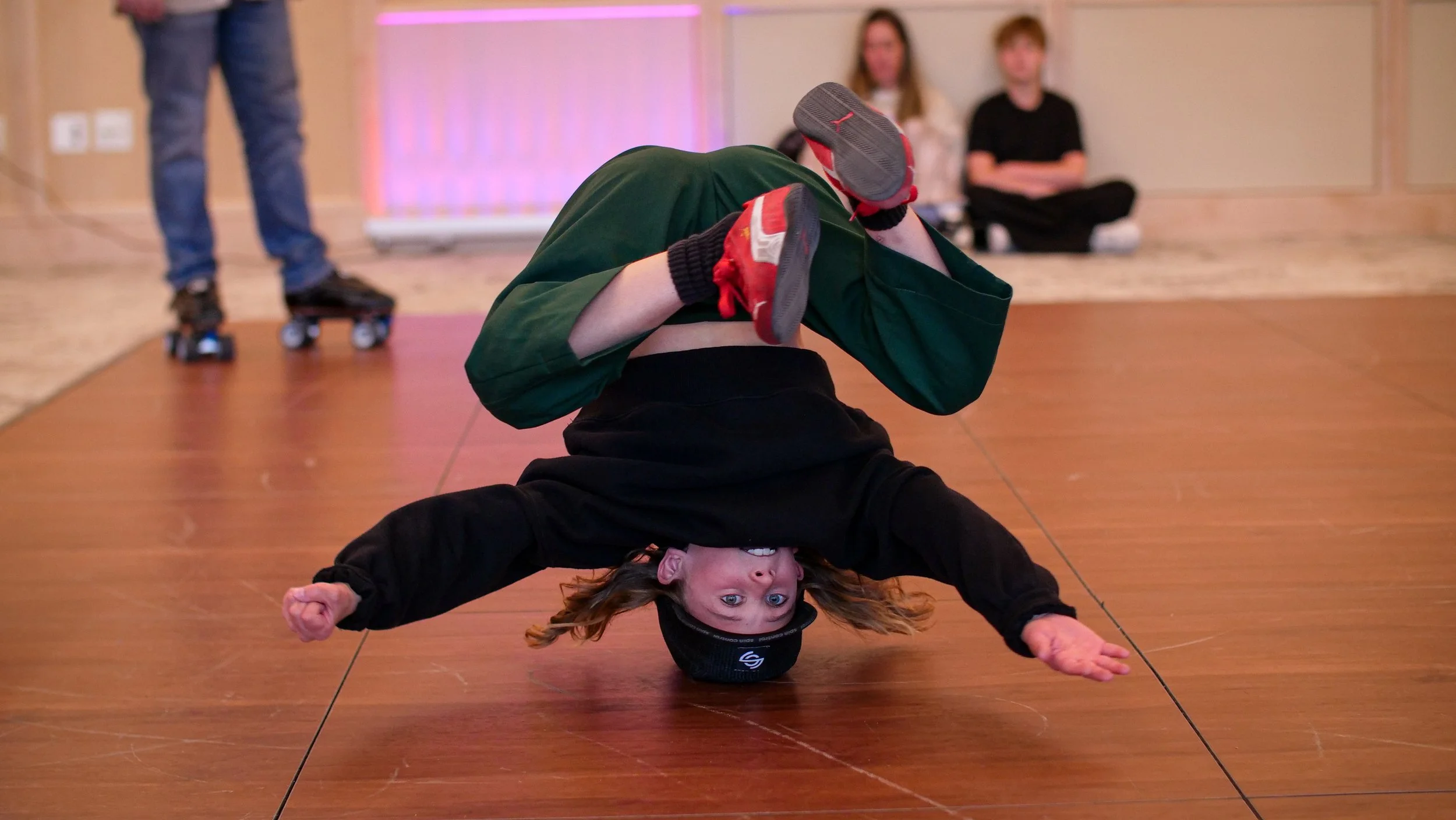 A girl performing an acrobatic pose with a person lying on the floor, both supporting each other with one arm each, in a room with other people in the background.