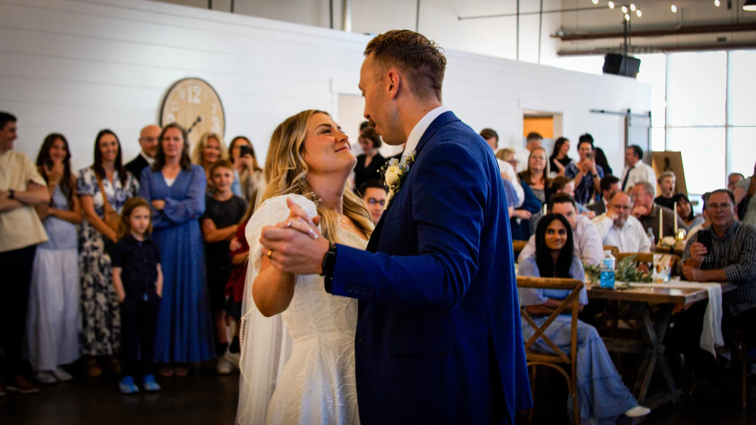 A bride and groom sharing a dance at their wedding reception with guests watching and smiling in the background.