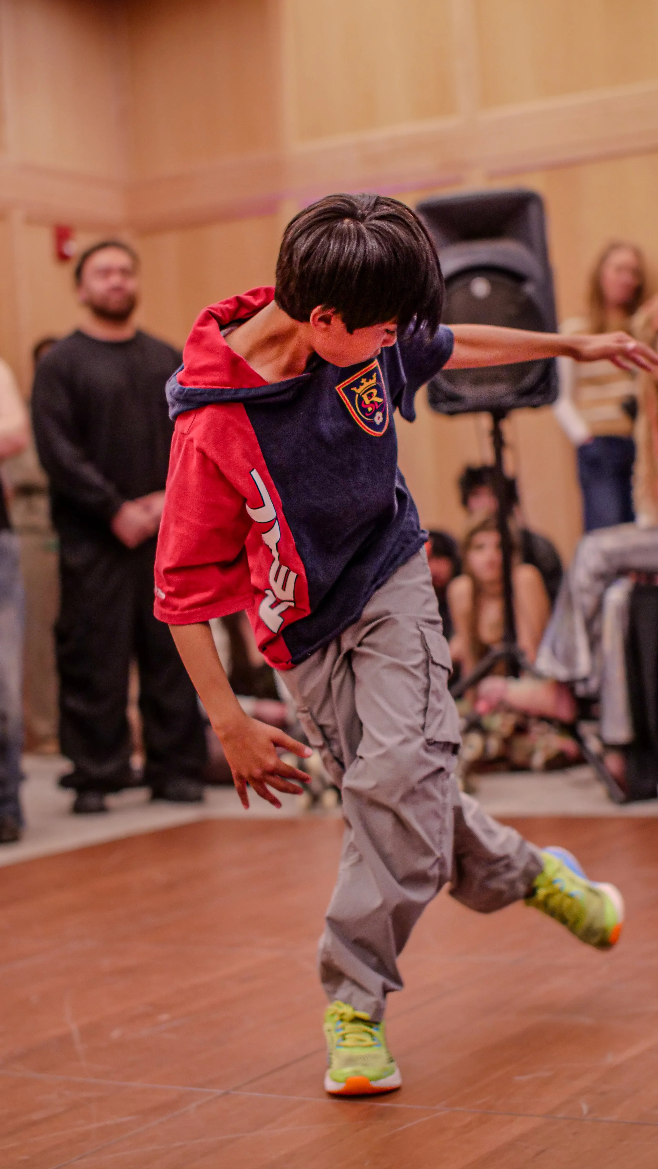 Young boy dancing or performing at an indoor event with onlookers in the background.