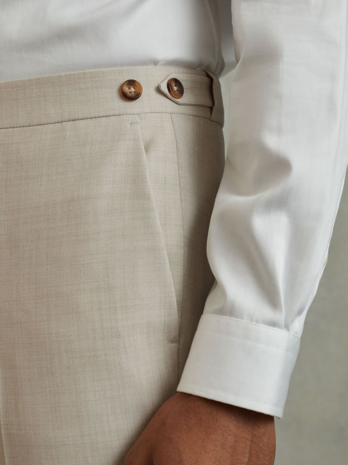 Close-up of a person wearing beige high-waisted trousers with brown buttons and a white long-sleeve shirt.