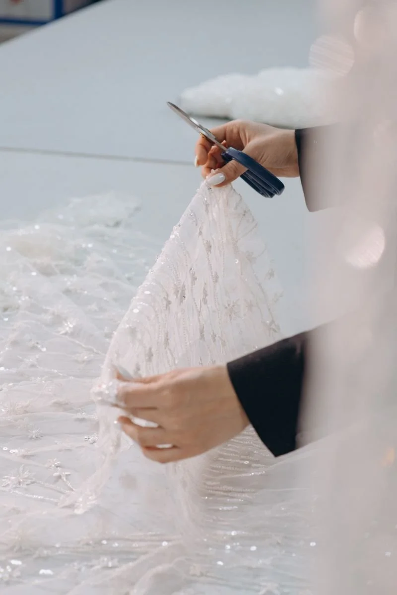 A person cutting lace fabric with scissors, likely preparing a wedding dress or gown.