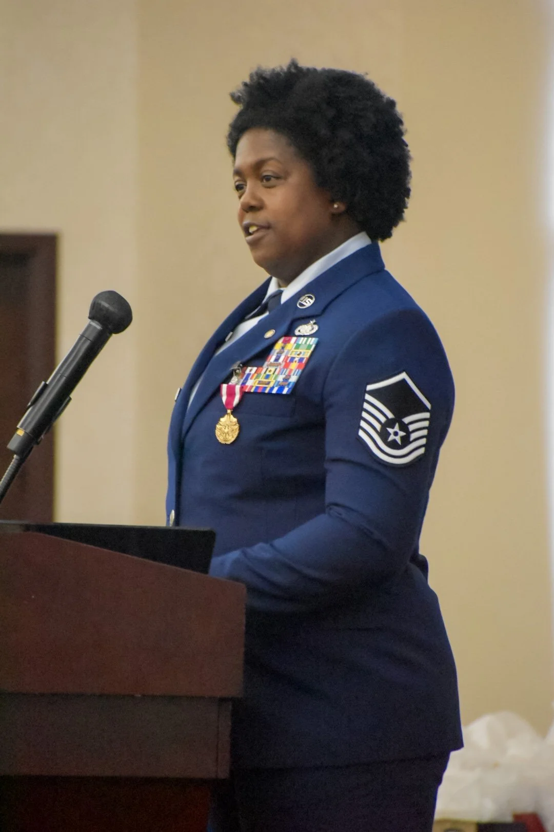A woman in a blue military uniform standing at a podium with a microphone, speaking at an event.