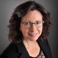 A woman with dark curly hair and glasses, smiling, wearing a black blazer and patterned blouse against a gray background.