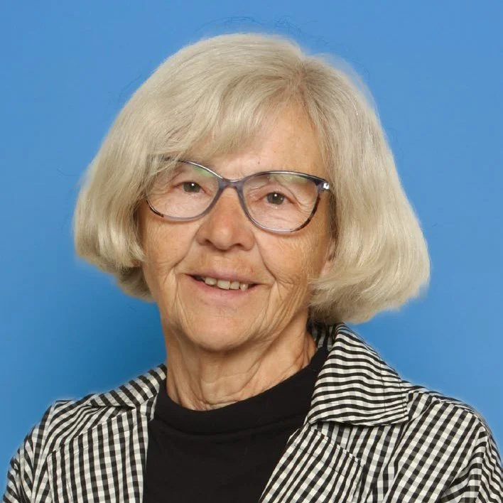 An elderly woman with light-colored hair, wearing glasses, a black top, and a black-and-white checkered shirt, smiling against a blue background.