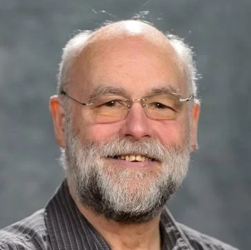 A smiling older man with glasses, a beard, and a bald head, wearing a checkered shirt.