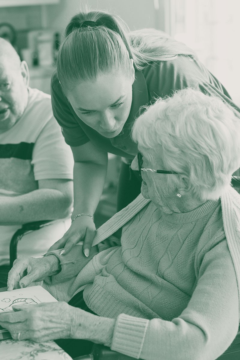 A caregiver leaning over an elderly woman with white hair, who is sitting and looking at a book, with another elderly man sitting nearby.