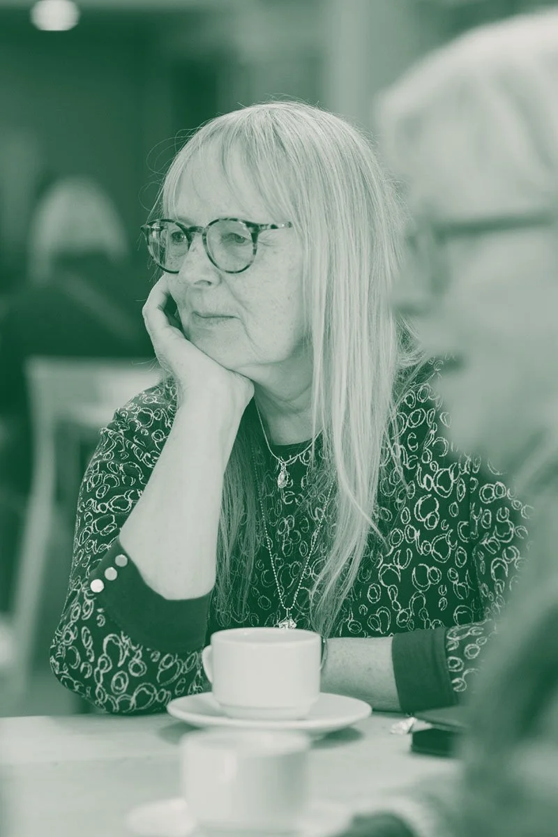 A woman sitting at a table with a cup, resting her chin on her hand, wearing glasses and a patterned shirt, in a cafe or restaurant setting.