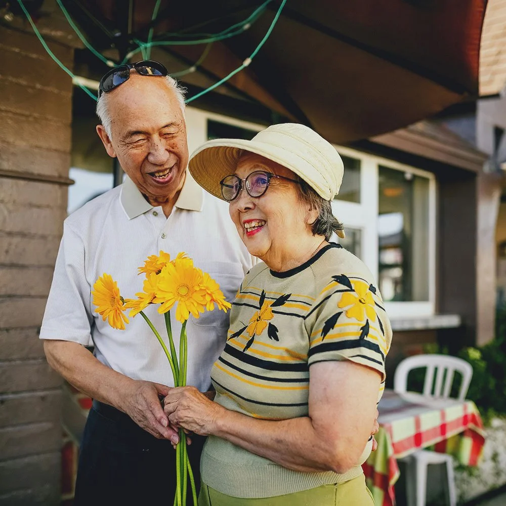 An elderly man and woman smiling, holding a bouquet of yellow flowers, outside a house with a striped tablecloth on a table in the background.