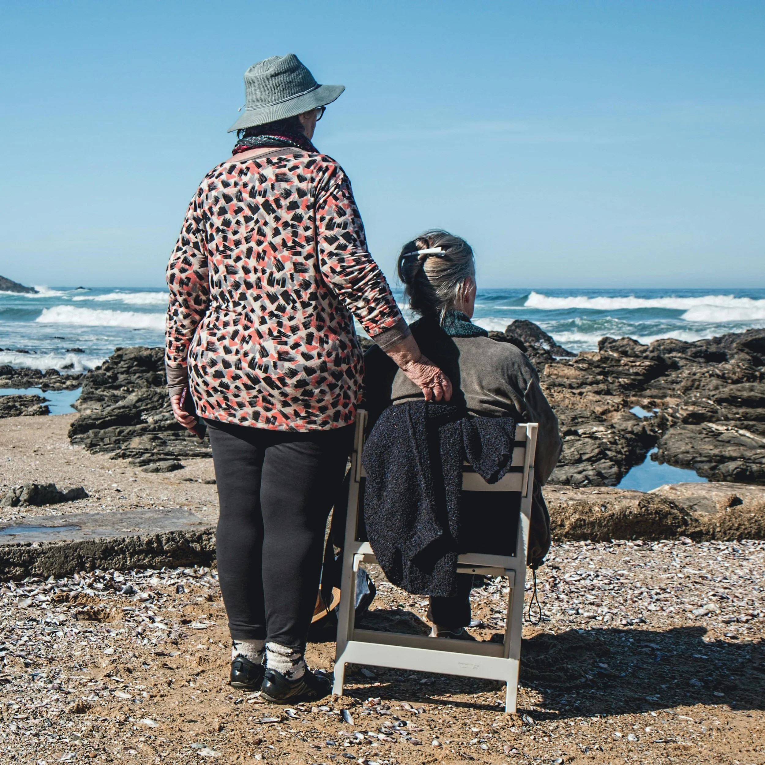 Two children and an adult at the beach with rocky shoreline and waves, one child sitting on a chair and the adult standing nearby, wearing a wide-brimmed hat and patterned jacket.