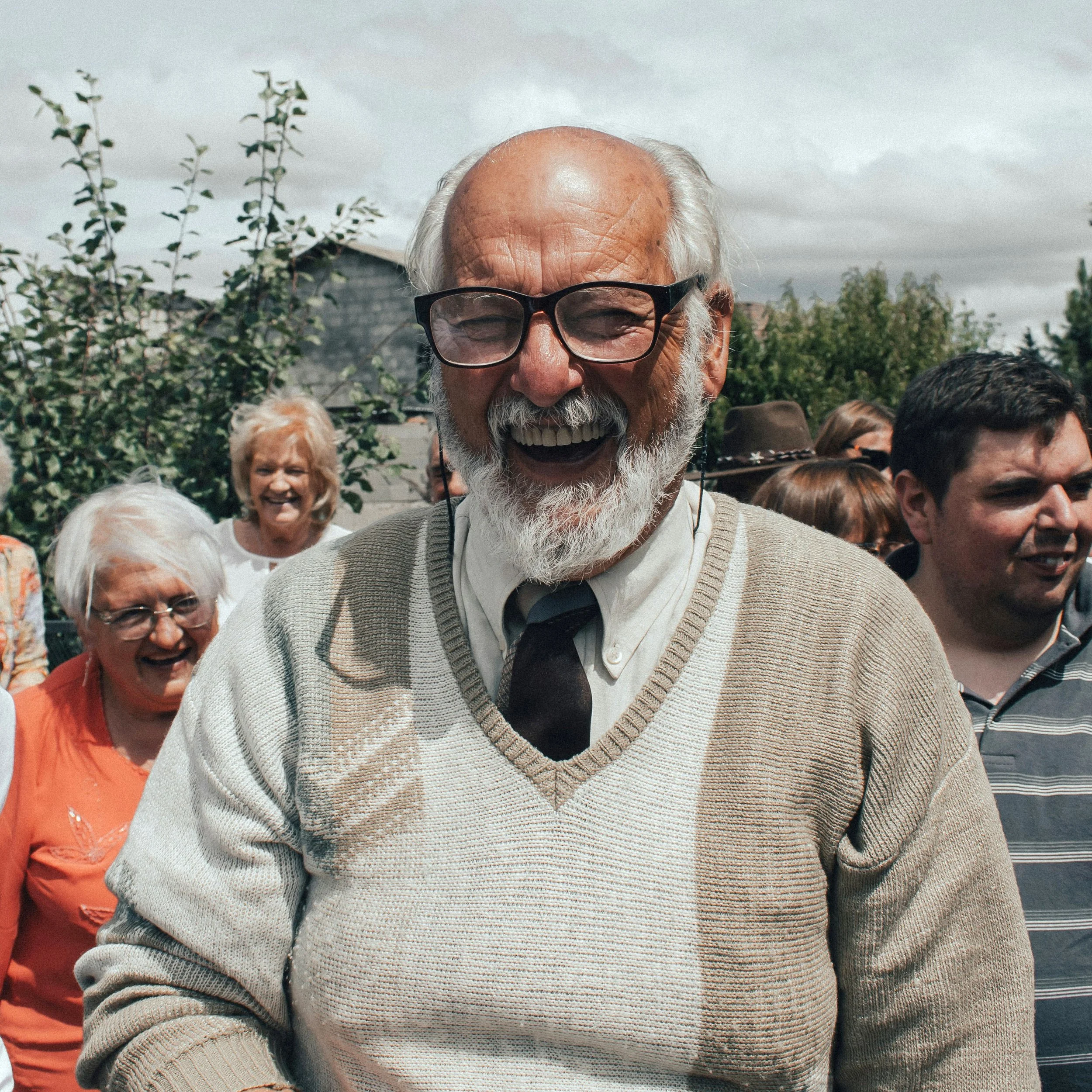 Close-up of an elderly man with glasses, white beard and bald head smiling at a gathering outdoors with cloudy sky, surrounded by other smiling people, including a woman in an orange top and others in casual attire.