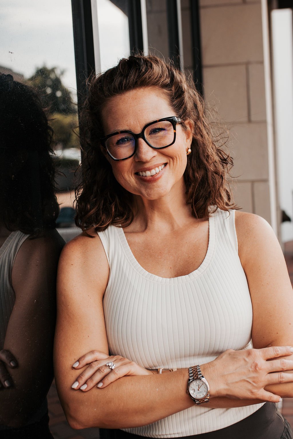 A woman with curly brown hair, wearing glasses and a white sleeveless top, smiling and standing next to a glass window.