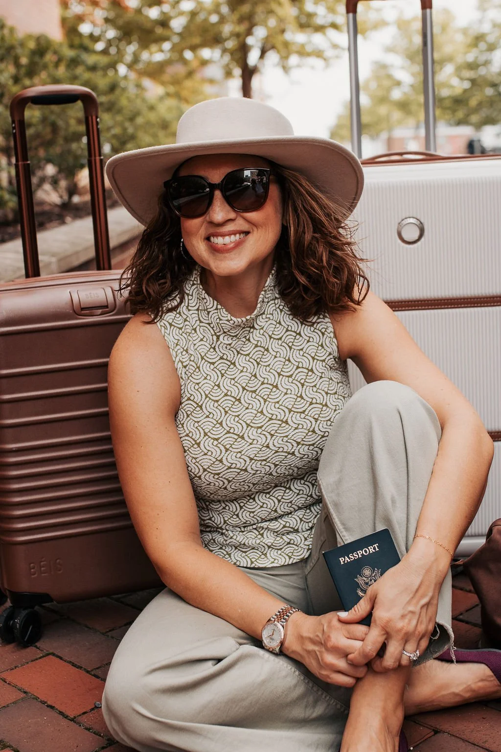 Woman sitting on brick ground with luggage and holding a passport, wearing a hat and sunglasses, smiling.