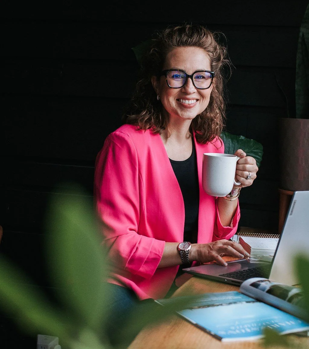 A woman smiling and holding a white coffee mug, sitting at a desk with a laptop and notebooks in a cozy indoor setting.