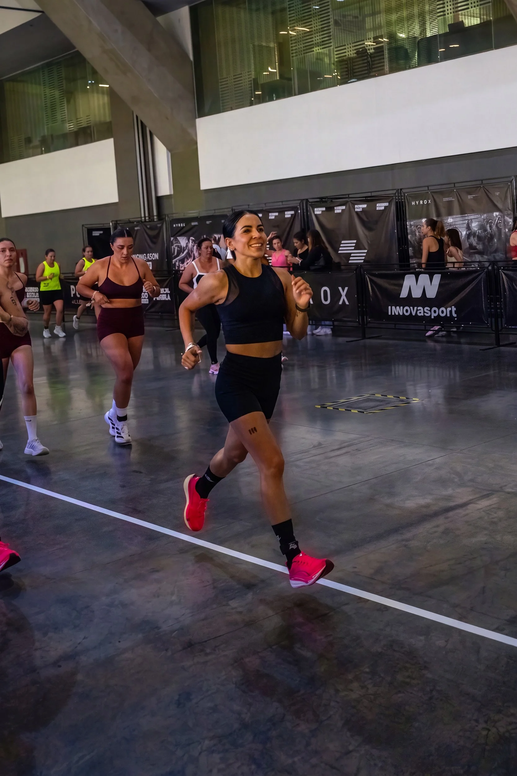 Mujer corriendo en competencia indoor con otras participantes, fotografía de evento fitness por Carlos Vega en Hyrox Puebla.