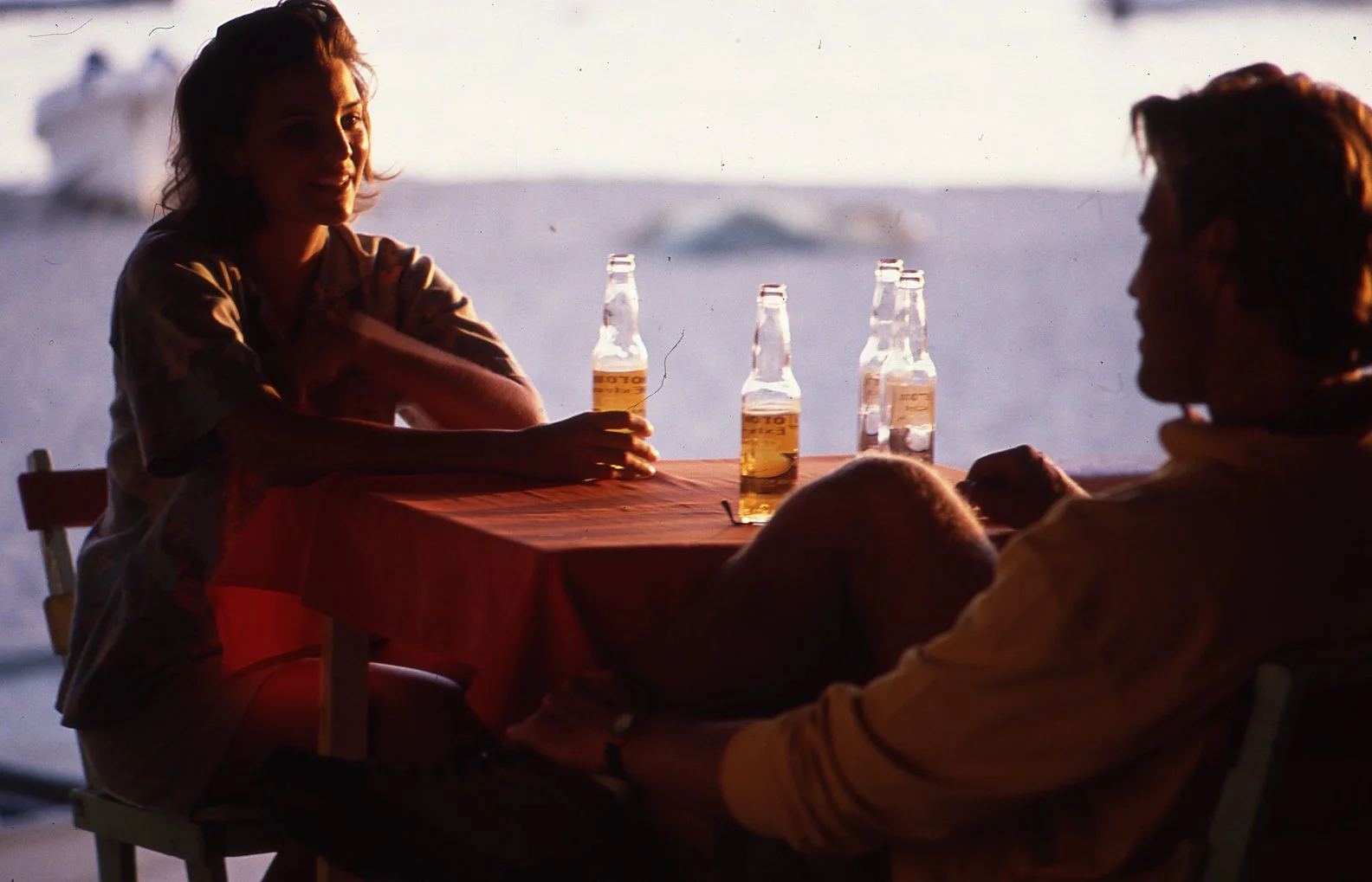 A woman and a man sitting at a table with four beer bottles, enjoying a drink outside at sunset near the sea.