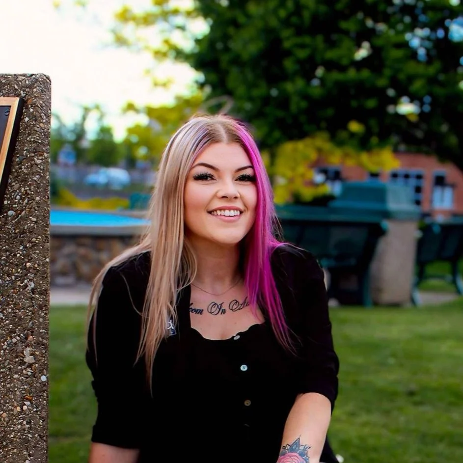 Young woman with blonde hair and pink streaks, smiling outdoors in a park with green trees and benches in the background.