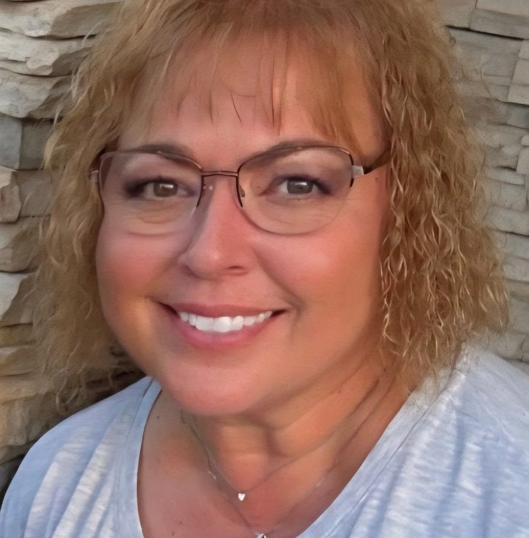 A smiling woman with curly blonde hair wearing glasses and a white top, against a background of a white stone wall.