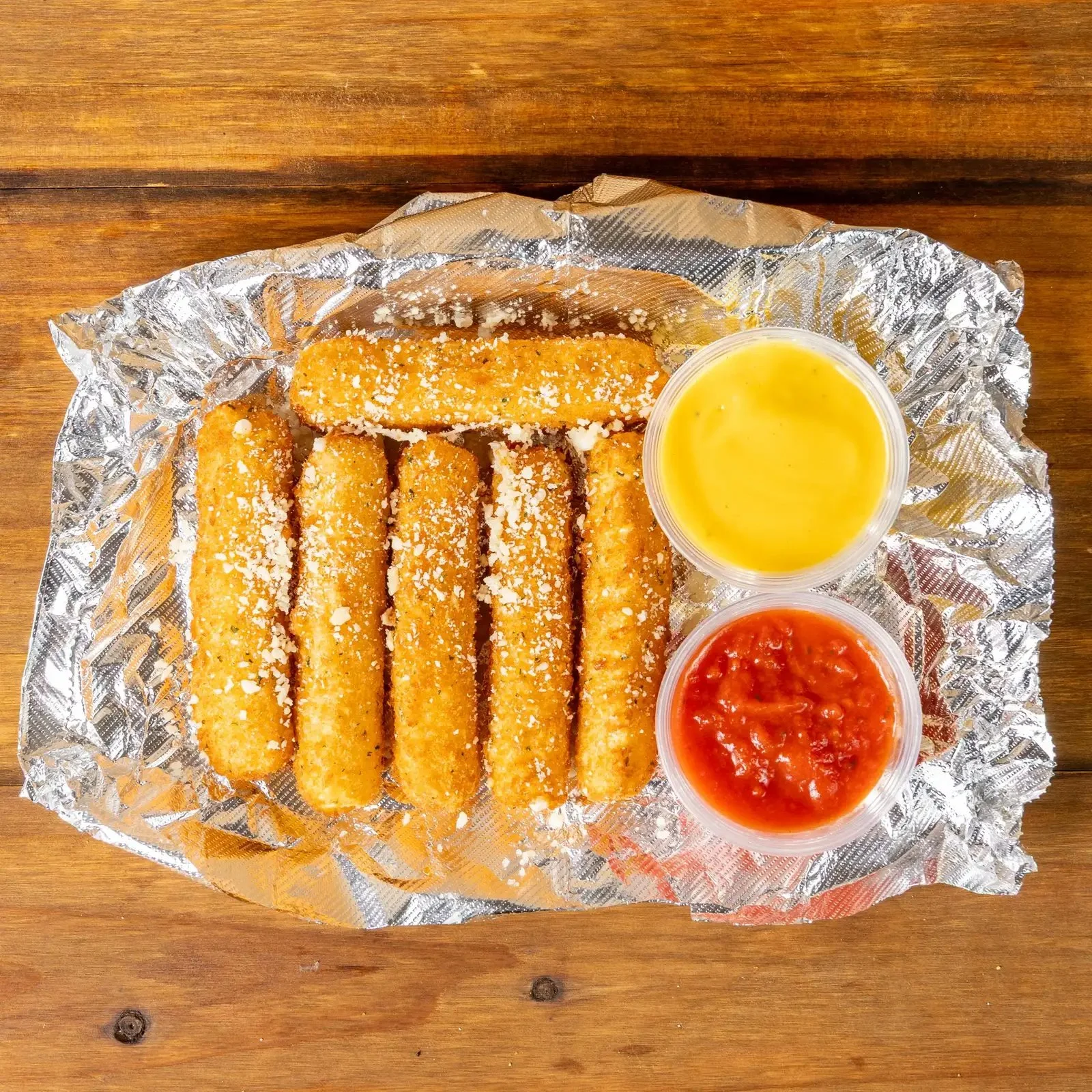 Crispy, breaded fish sticks topped with grated cheese, served with two cups of dipping sauces, one yellow and one red, on a sheet of aluminum foil on a wooden surface.