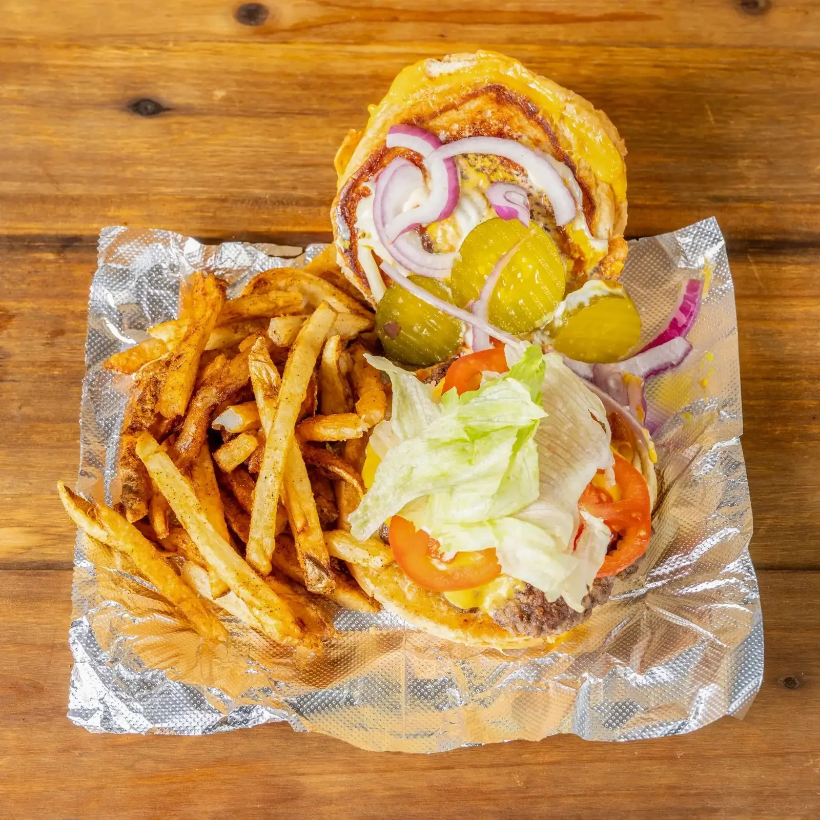 A cheeseburger with pickles, onions, lettuce, tomato in a bun, served with French fries on a piece of aluminum foil on a wooden table.