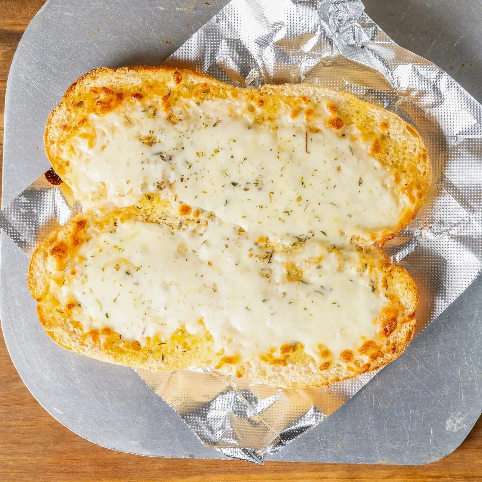 Slices of garlic bread topped with melted cheese and herbs placed on aluminum foil on a round metal tray.