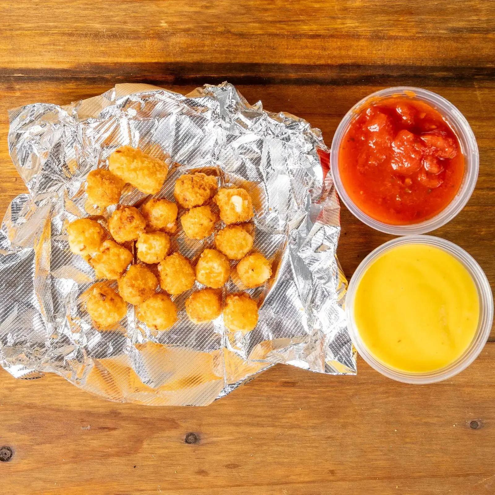 Basket of fried cheese balls with two containers of dipping sauces, one red and one yellow, on a wooden table.