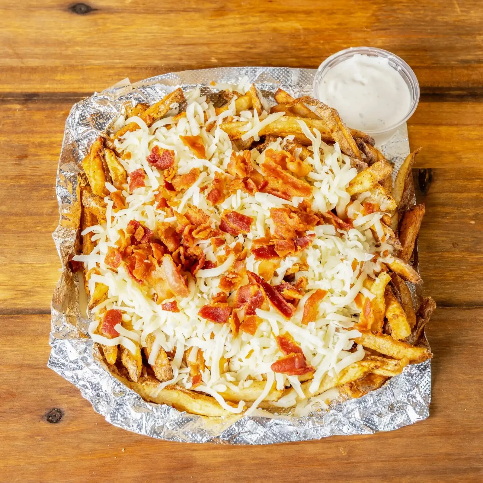 Fried potato fries topped with shredded cheese and crispy bacon bits, served with a side of ranch dipping sauce on a wooden table.