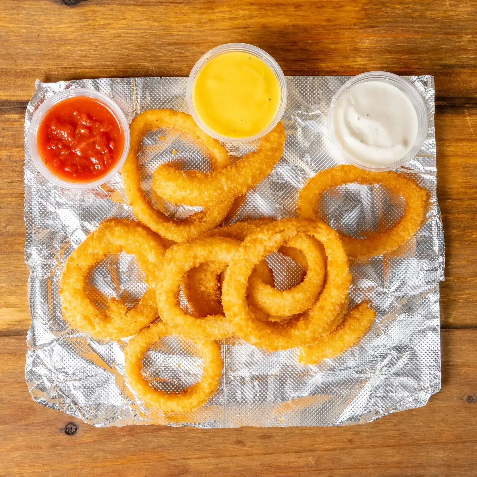Golden-brown onion rings on aluminum foil with three dipping sauces: red ketchup, yellow cheese, and white ranch, on a wooden surface.