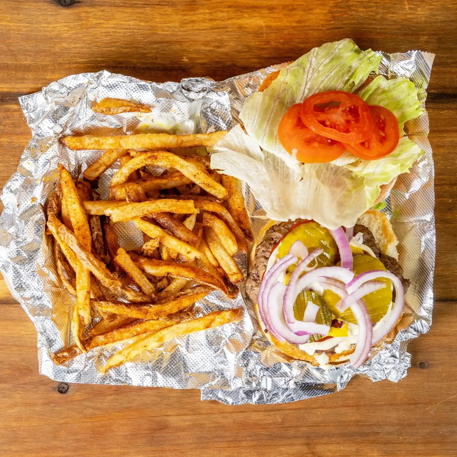 A cheeseburger with lettuce and tomato, served with seasoned French fries and pickles with onions on a sheet of foil on a wooden surface.