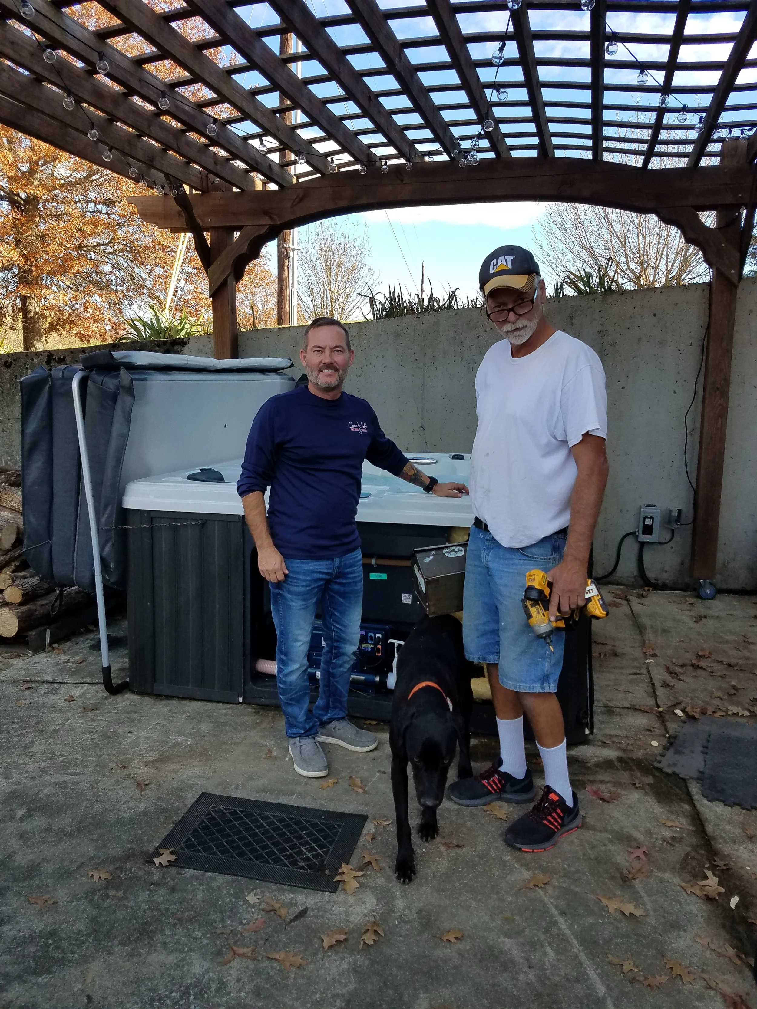 Two men standing outdoors next to a hot tub with a dog in front of them, holding two cordless drills. One man is wearing a white t-shirt, shorts, and a cap, while the other is wearing a dark long-sleeve shirt and jeans. They are under a wooden pergol