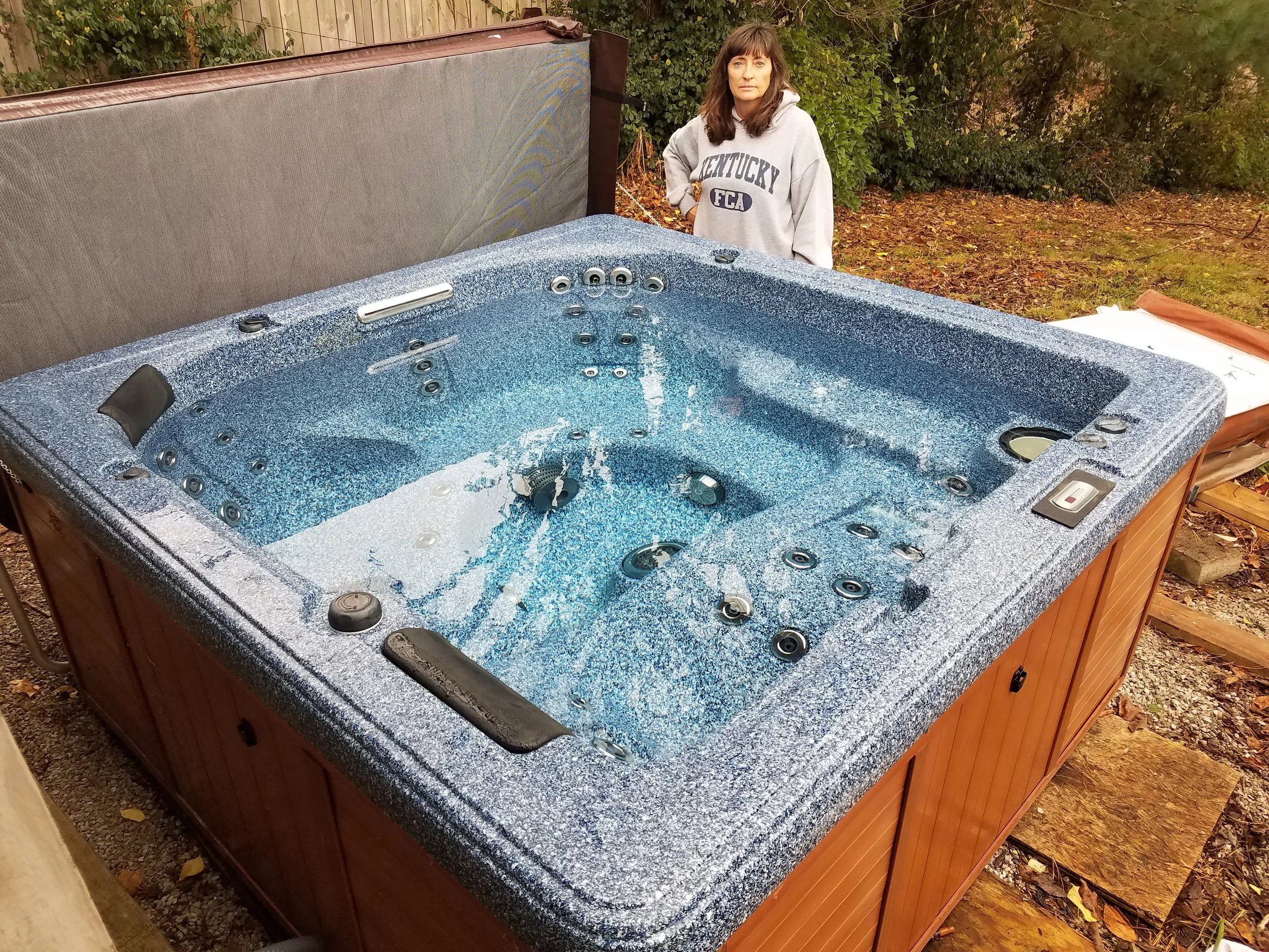 A woman with brown hair wearing a gray sweatshirt that says Kentucky FCA stands beside an outdoor hot tub filled with water, in a backyard with trees and bushes, fall leaves on the ground.