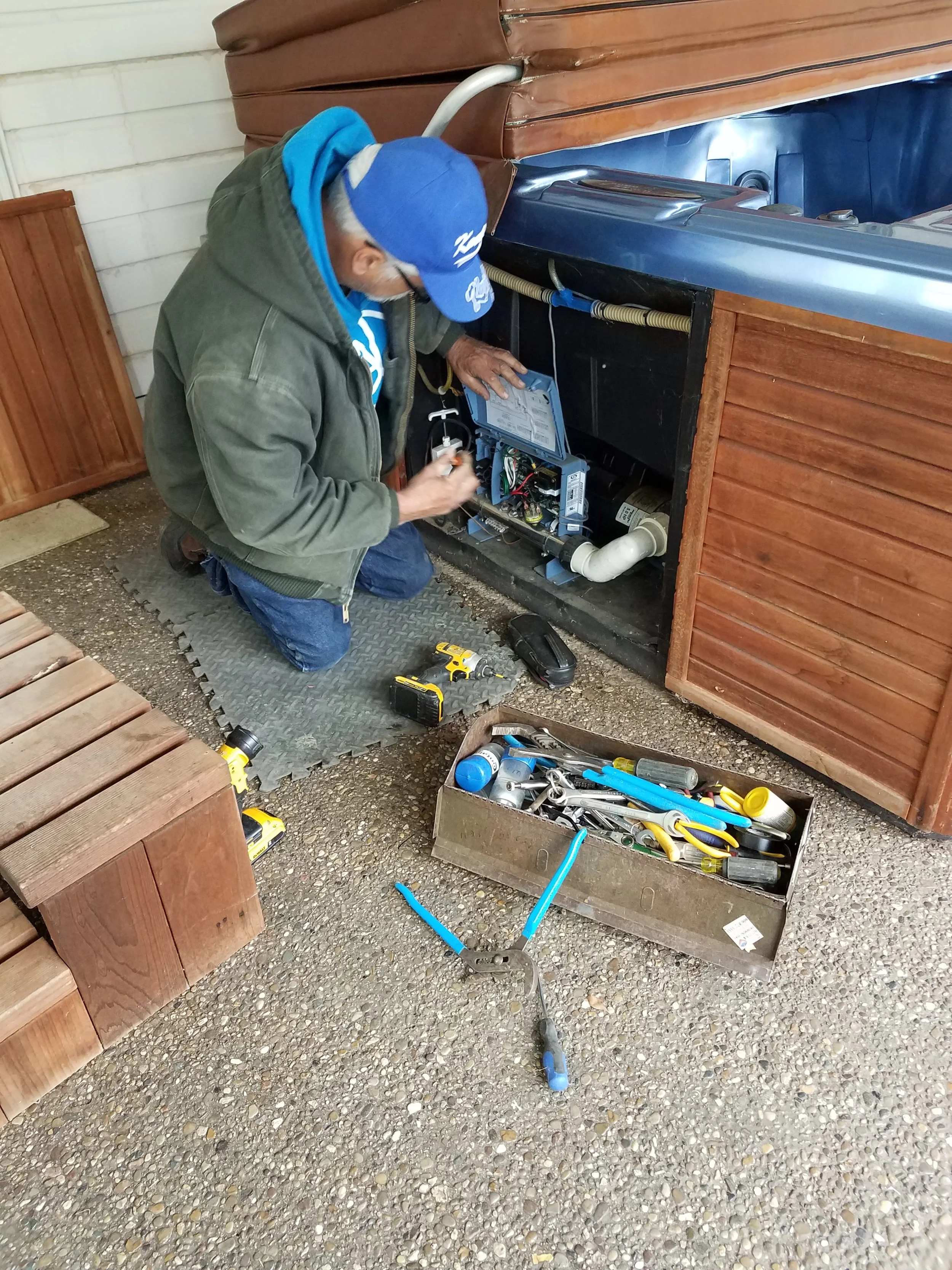 A person working on an open electrical panel or control box, kneeling on a rubber mat, surrounded by tools including a cordless drill, pliers, and screwdrivers, with a table of tools nearby.