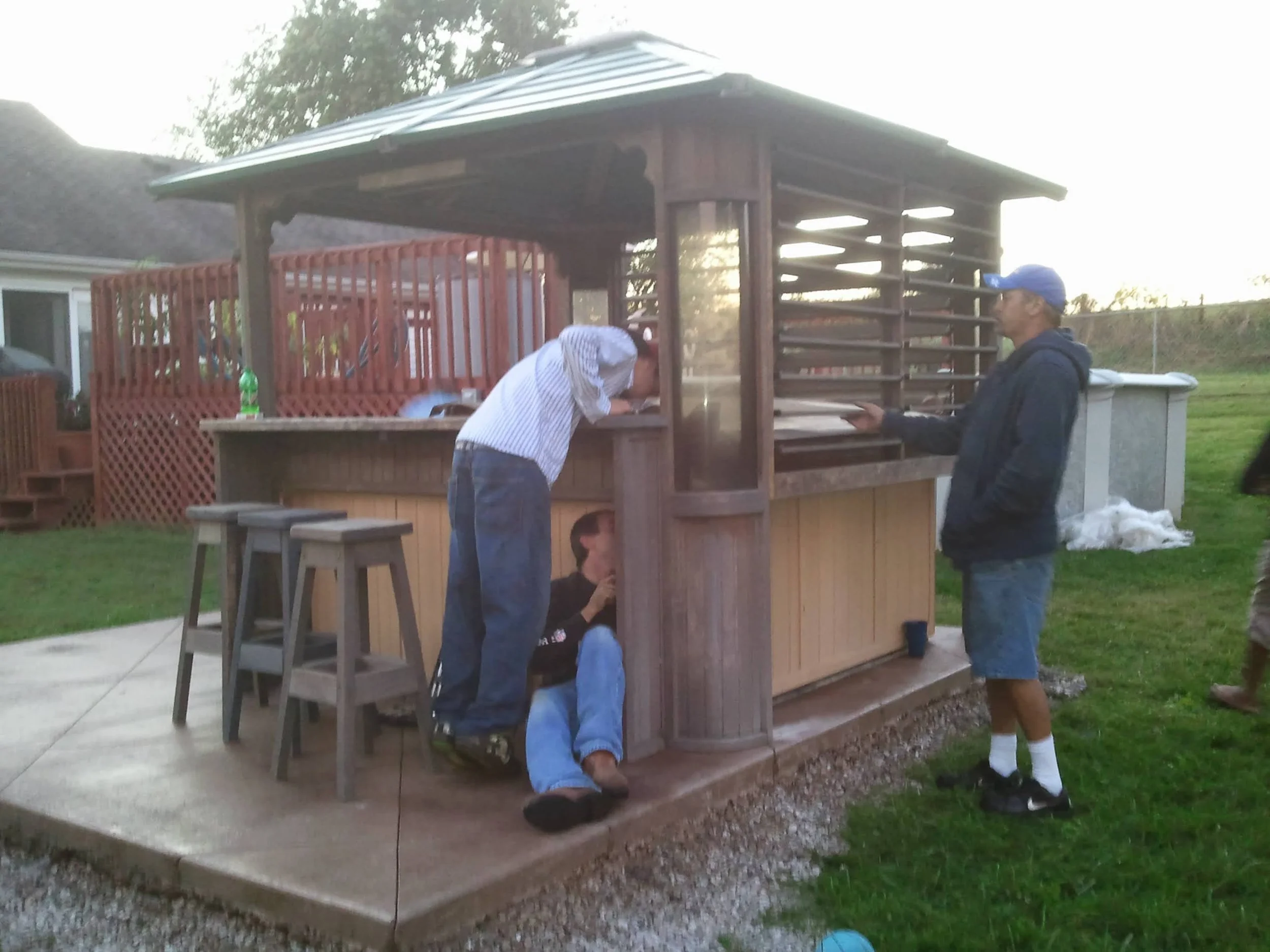 Three children working on building a wooden outdoor bar or kiosk in a backyard, with a metal roof. One child is leaning over the counter, another under the counter, and a third standing nearby holding tools.