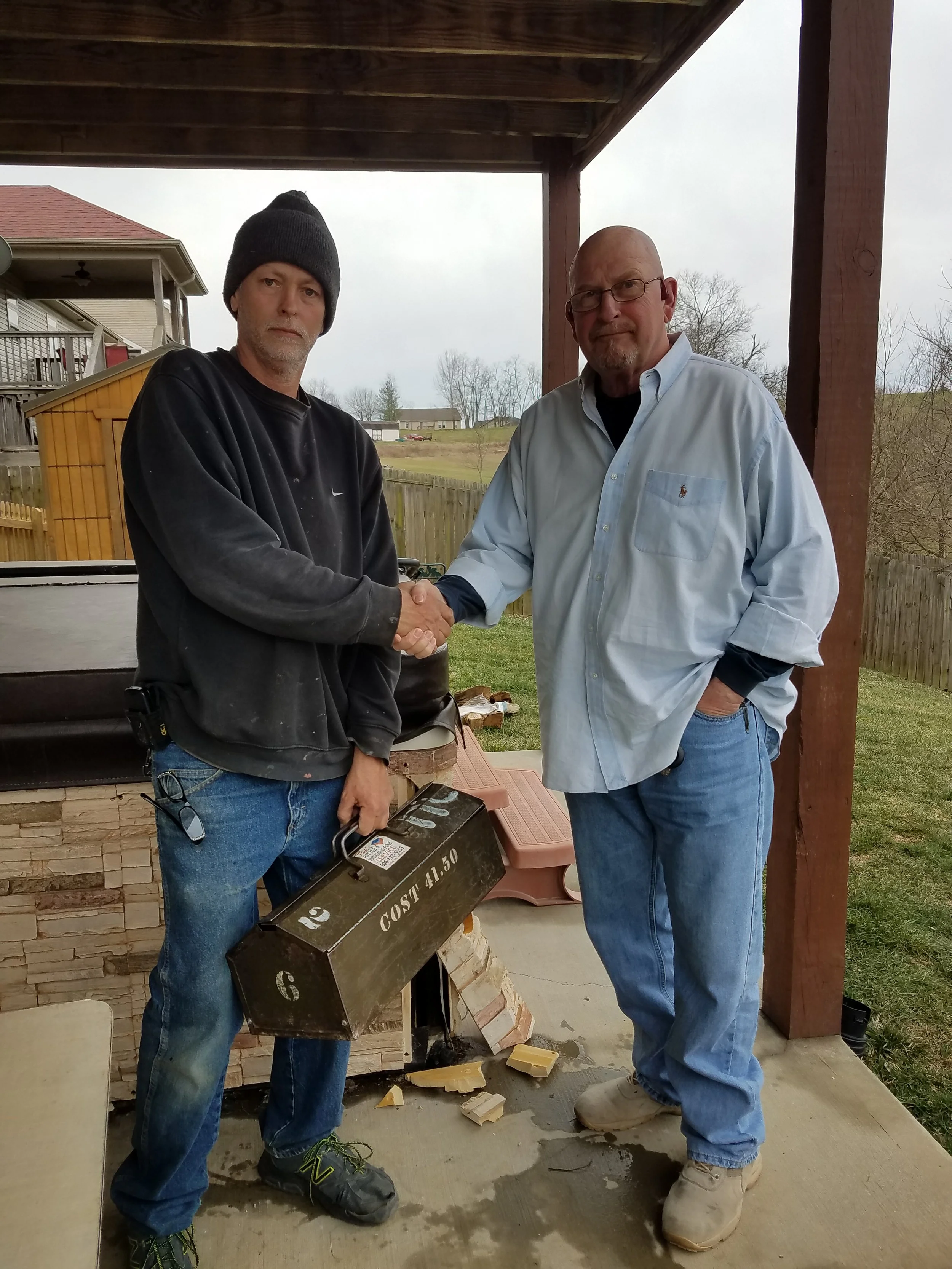 Two men shaking hands outdoors under a wooden porch, one in a black sweatshirt and beanie, the other in a light blue shirt and glasses, with a cloudy sky and residential area in the background.