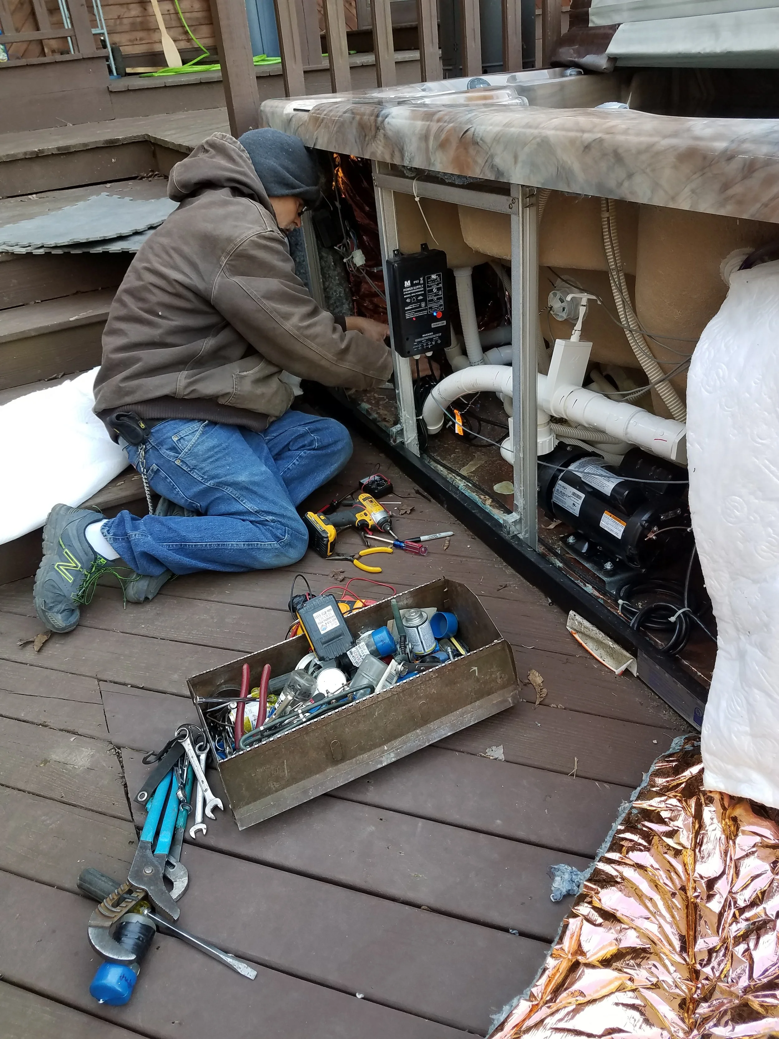 A person working on an under-sink plumbing system outside on a wooden deck. The person is wearing a hoodie, glasses, and jeans, surrounded by tools and pipes, with pipe insulation and foam around the area.