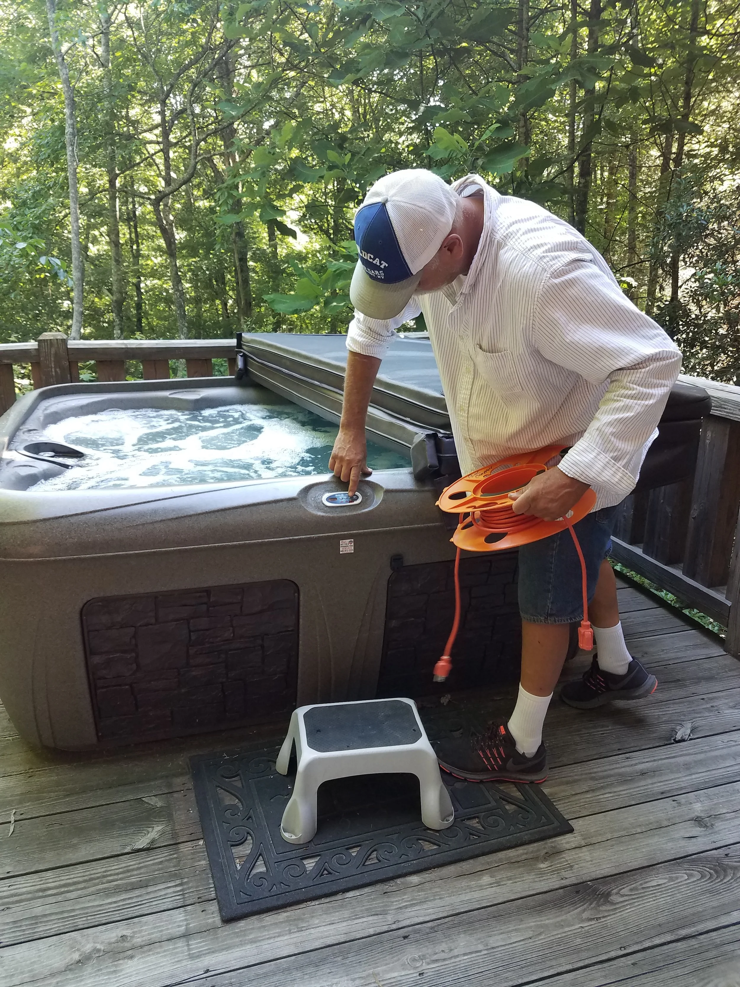 A man in a striped shirt and cap checks a hot tub on a wooden deck surrounded by trees. He holds an extension cord and points to a control panel on the hot tub.