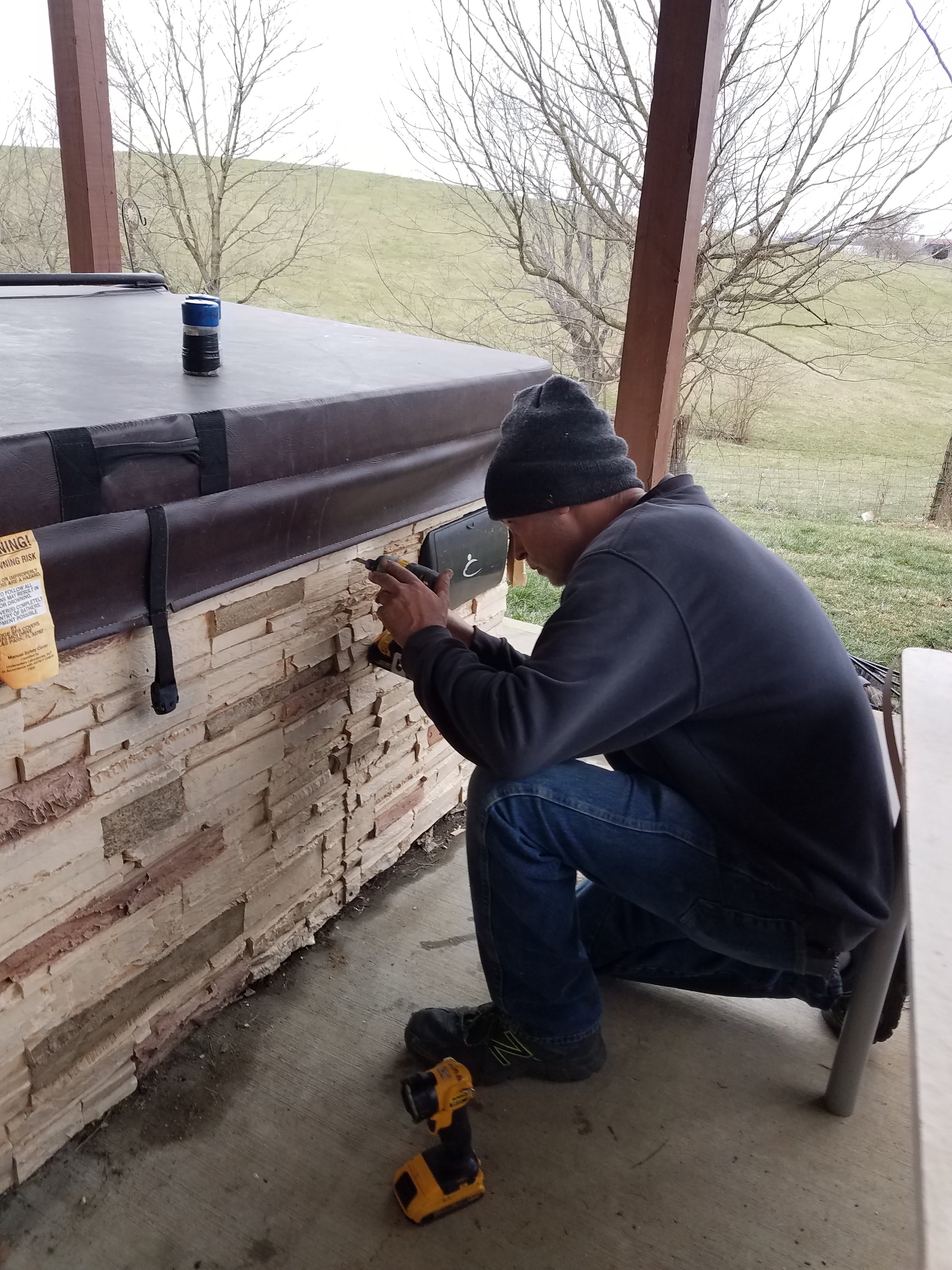 Man wearing a black beanie and a dark jacket kneeling on a concrete floor using a cordless drill on a stacked stone wall. A yellow cordless drill lies on the ground near him. The scene appears to be outdoors under a wooden porch or deck with a grassy yard and leafless trees in the background.