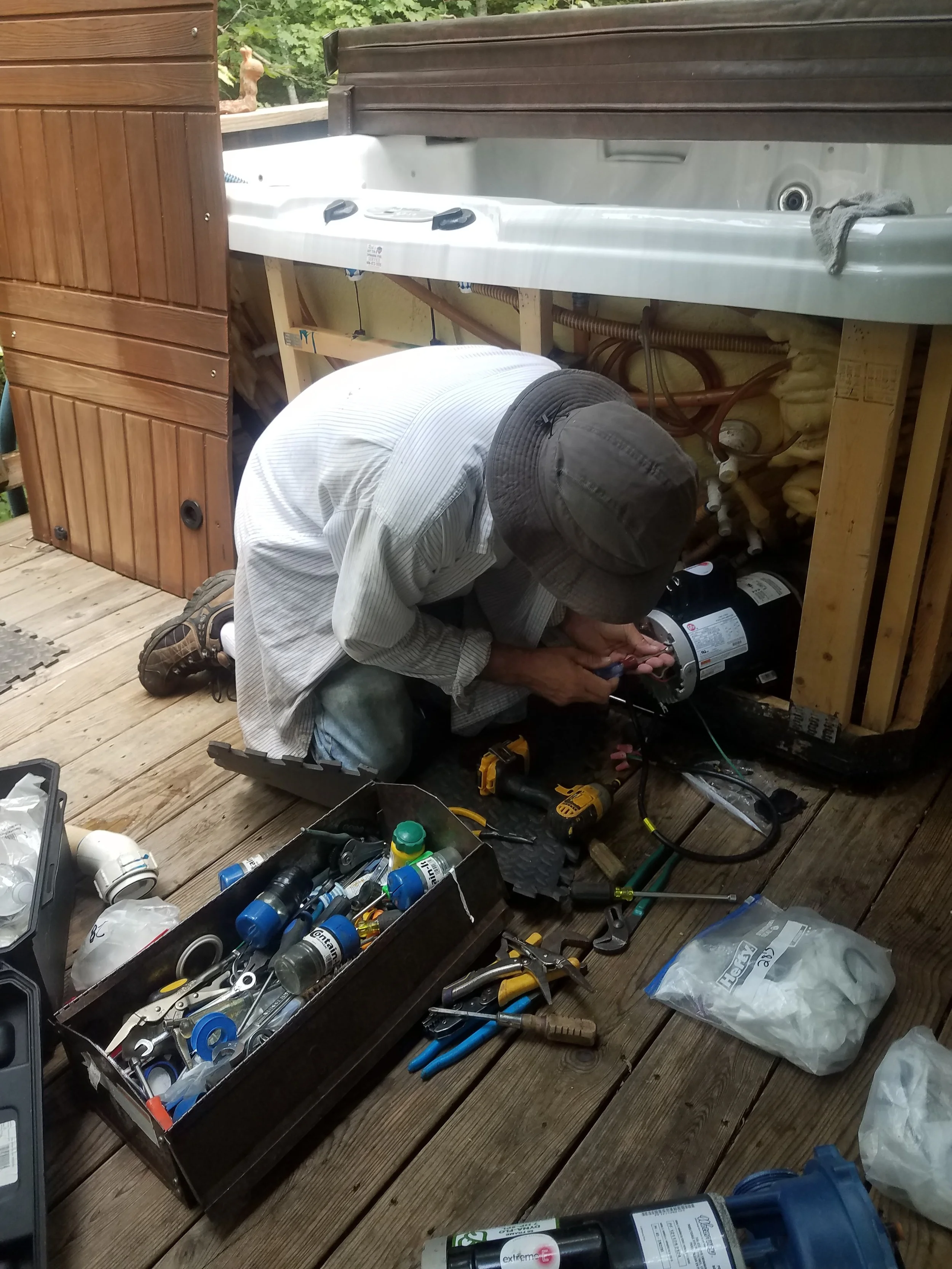 A person working on installing or repairing a hot tub outside on a wooden deck, surrounded by tools and supplies.